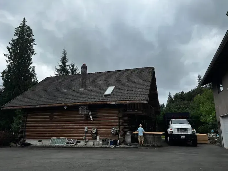 Log cabin with a person and parked van under a cloudy sky, surrounded by trees