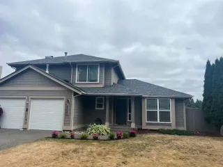 Suburban two-story gray house with white garage doors, front porch, and dry yard under cloudy sky