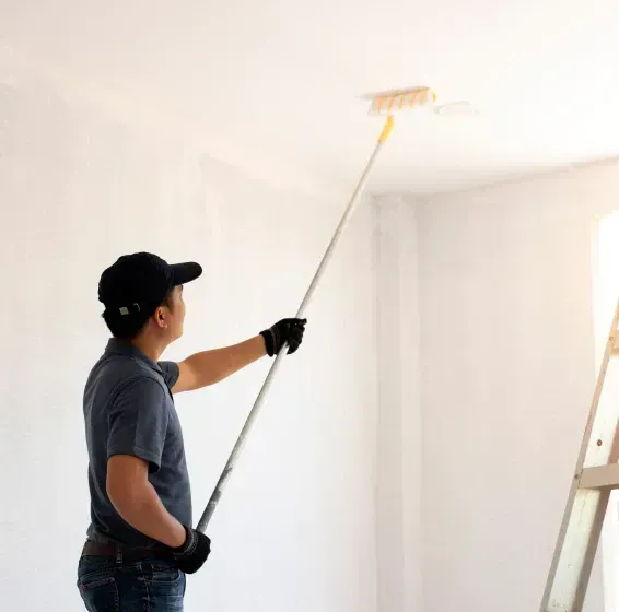Person painting a white ceiling with a roller in a bright room, standing by a ladder.