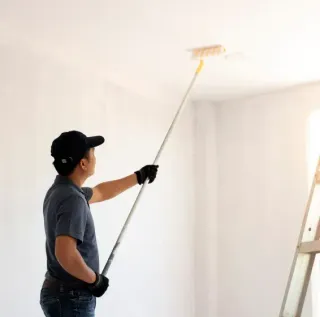 Person painting a white ceiling with a roller in a bright room, standing by a ladder.