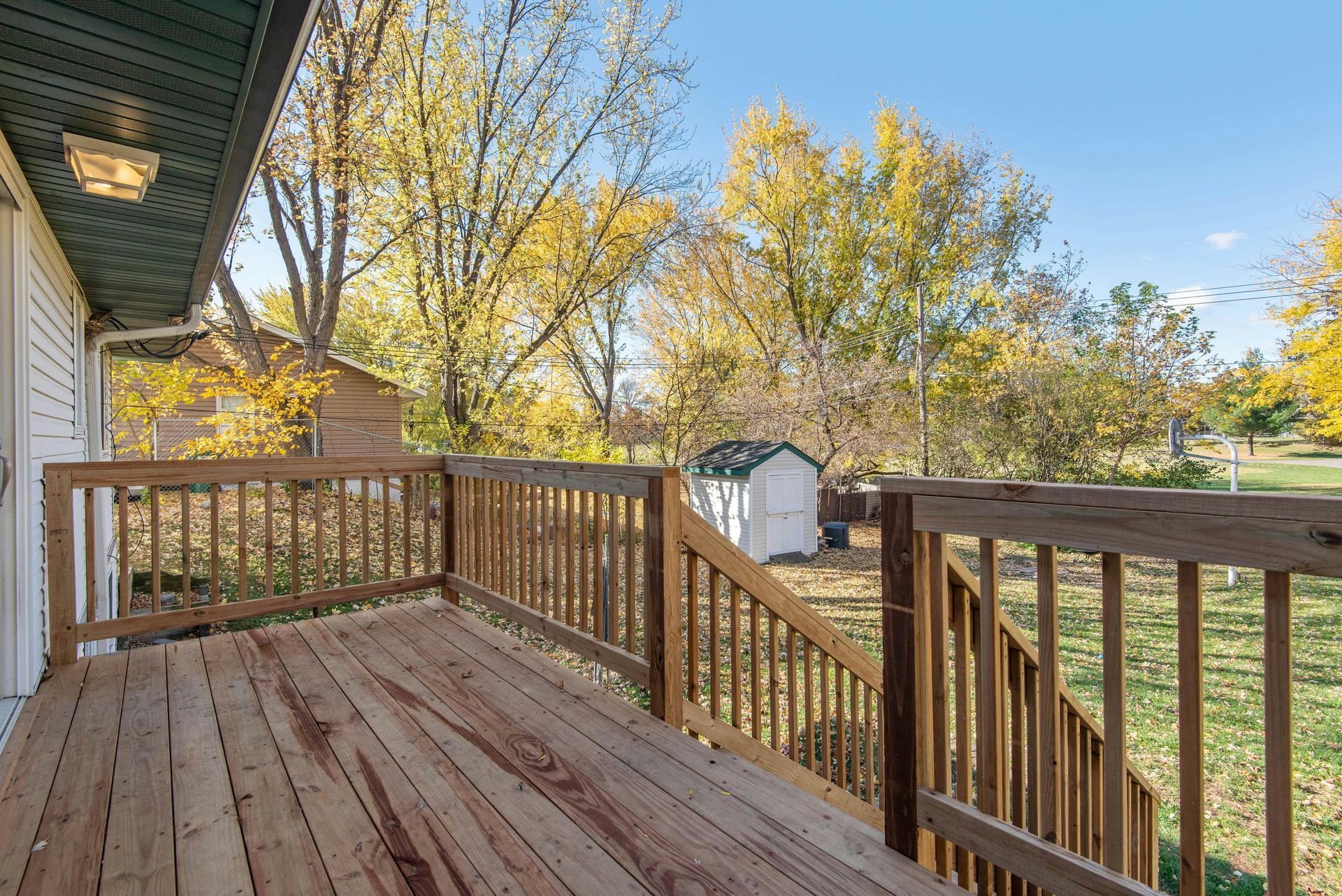 Wooden deck with railing beside a house, overlooking a grassy yard and autumn trees under a blue sky