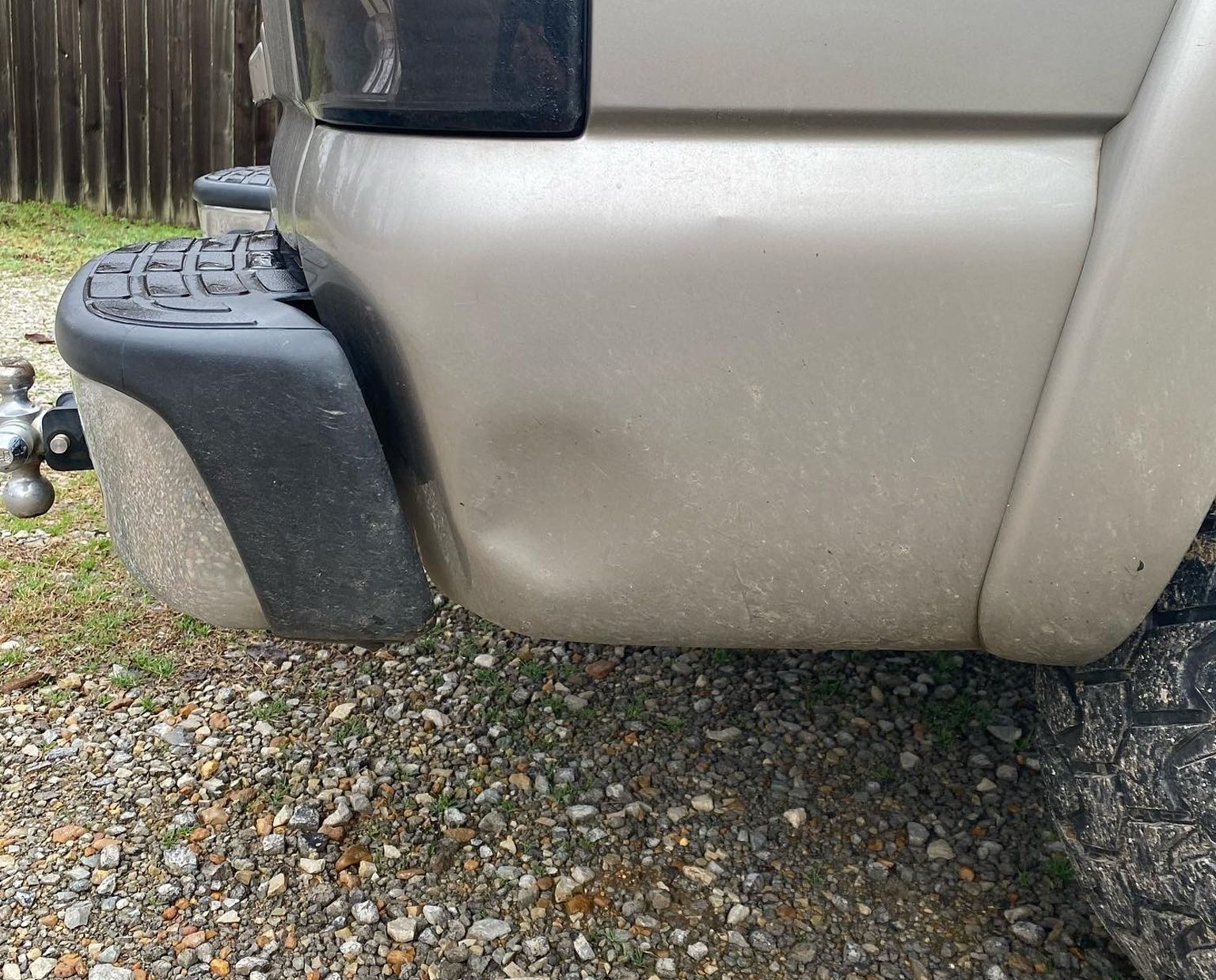 Close-up of a beige vehicle’s rear corner, showing a black step bumper and gravel ground.