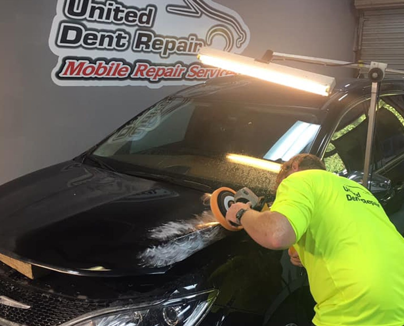 Technician polishing a black car hood in a mobile dent repair shop under bright work lights