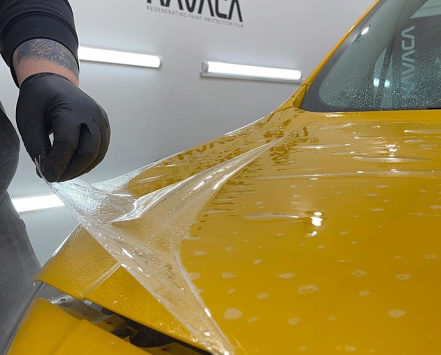 Gloved hand peeling clear film from a yellow car hood in a detailing shop