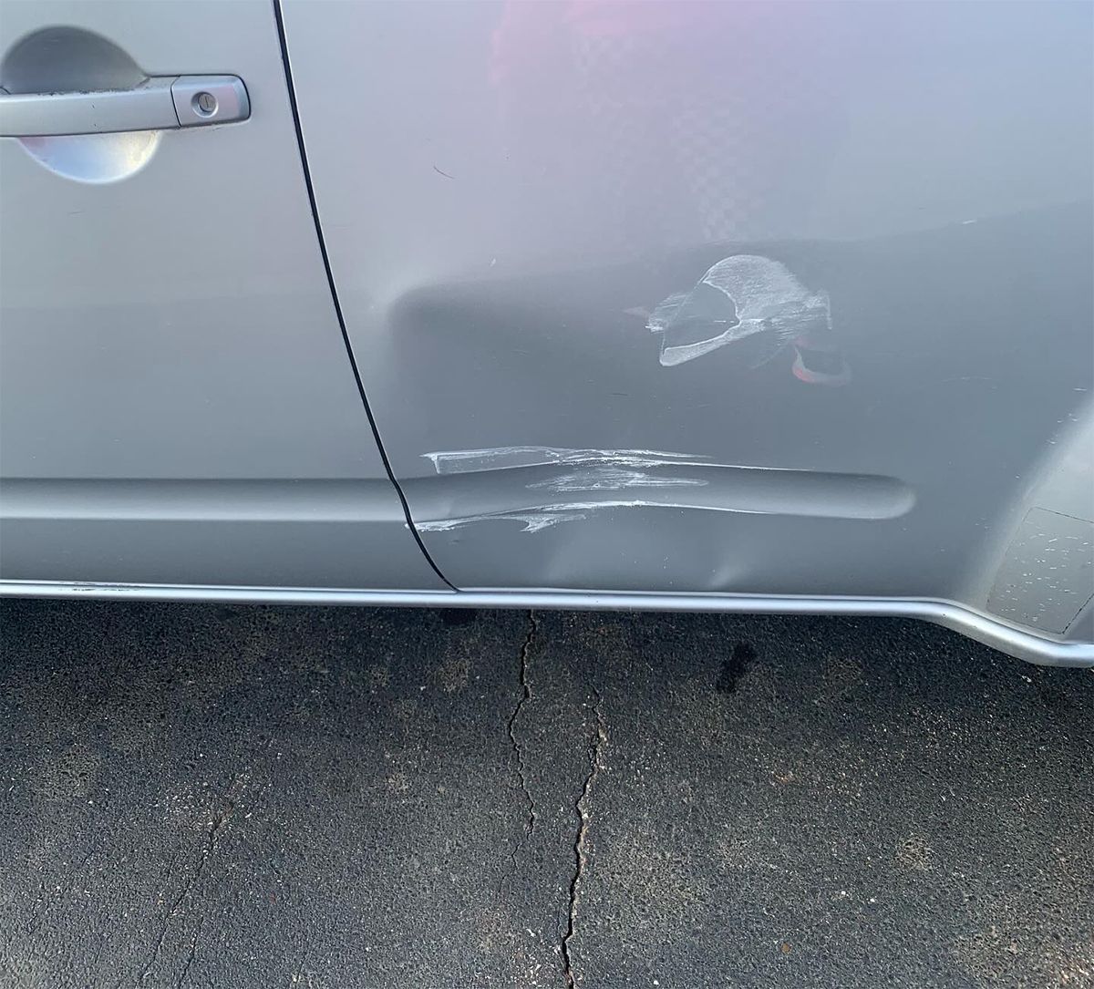 Silver car door with dents and white scrape marks near the bottom on a paved surface