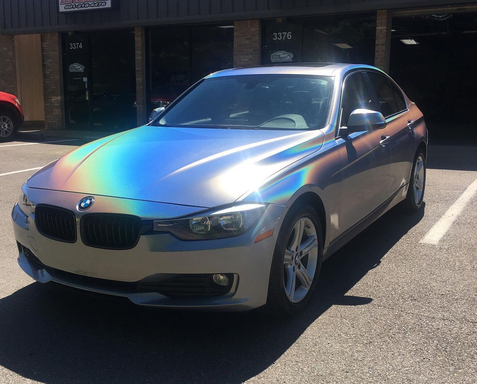 Silver BMW sedan parked in sunlight outside a storefront, with a rainbow reflection on the hood.