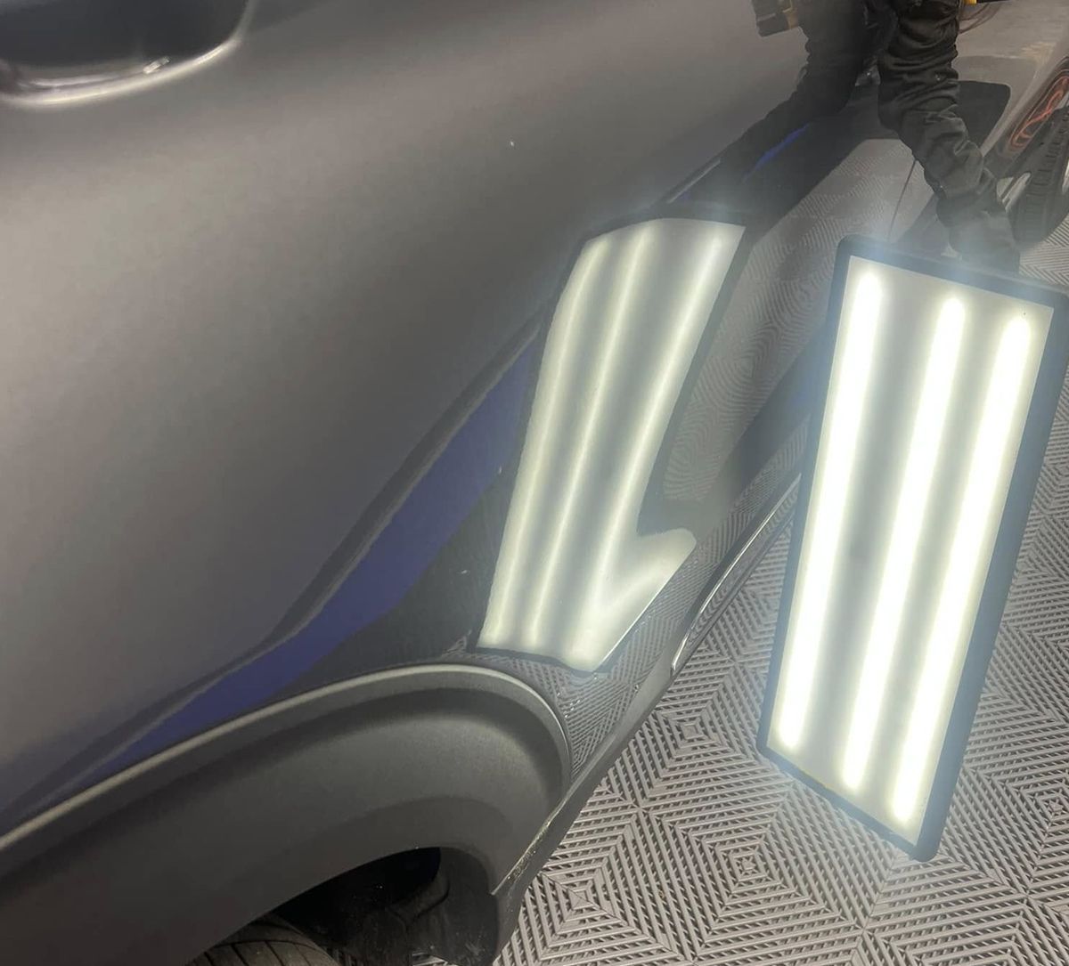 Close-up of a dark car door reflecting bright overhead lights on a patterned floor.