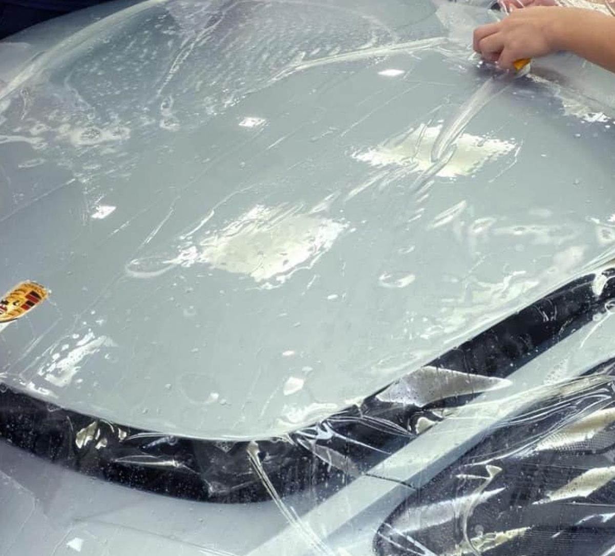A hand peeling clear plastic film from a glossy gray car surface inside a garage.