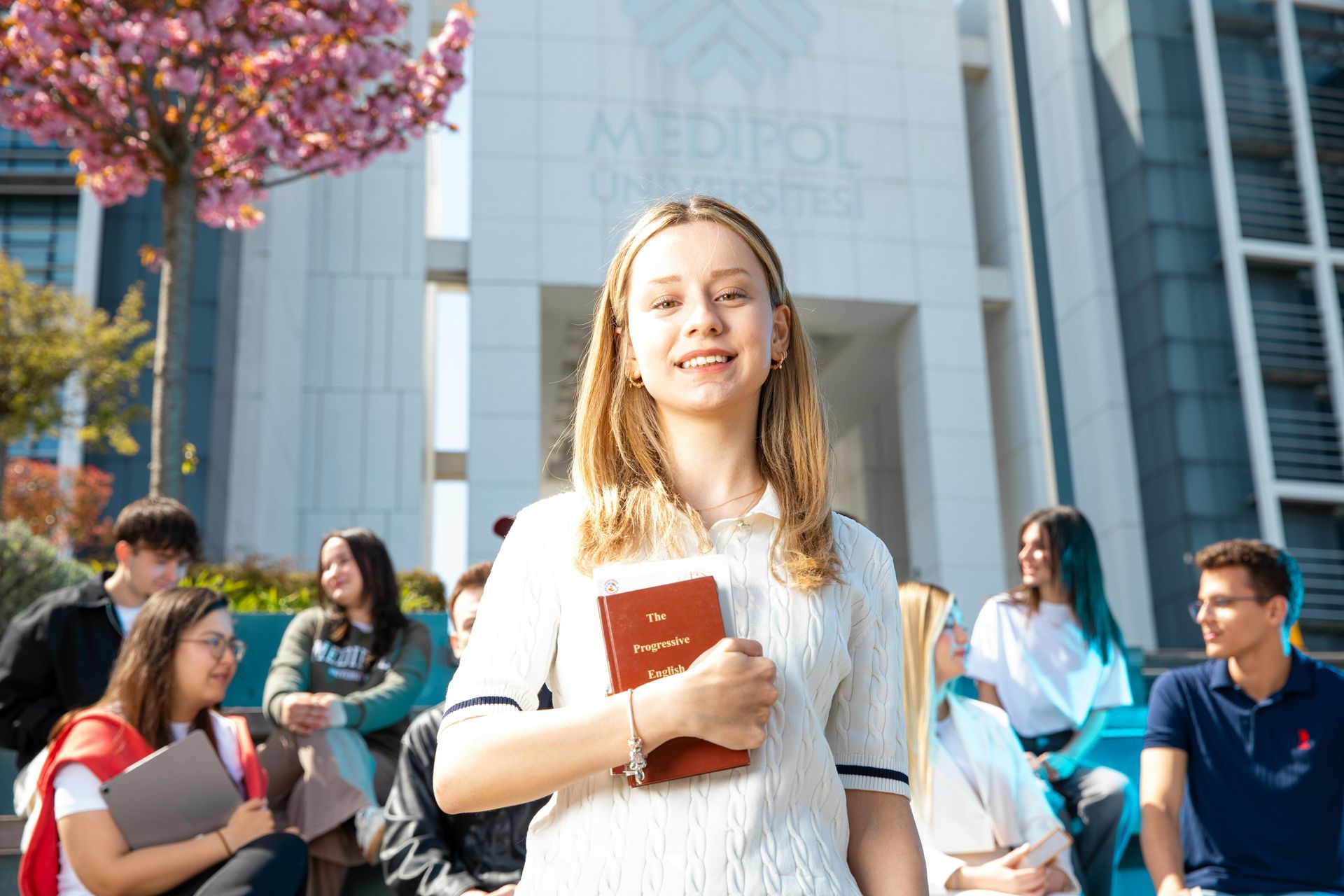 Una giovane donna sorride con in mano un libro davanti a un edificio universitario; sullo sfondo ci sono degli studenti.