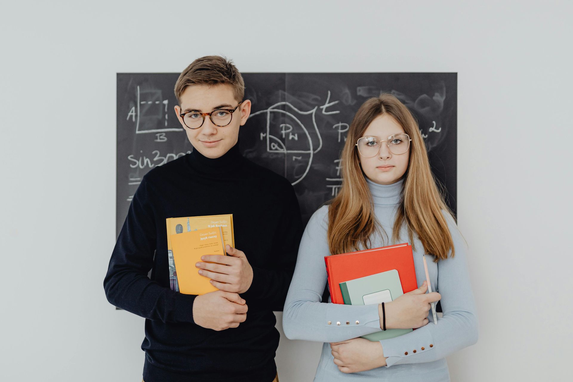 Due persone con occhiali e libri davanti a una lavagna con equazioni matematiche.