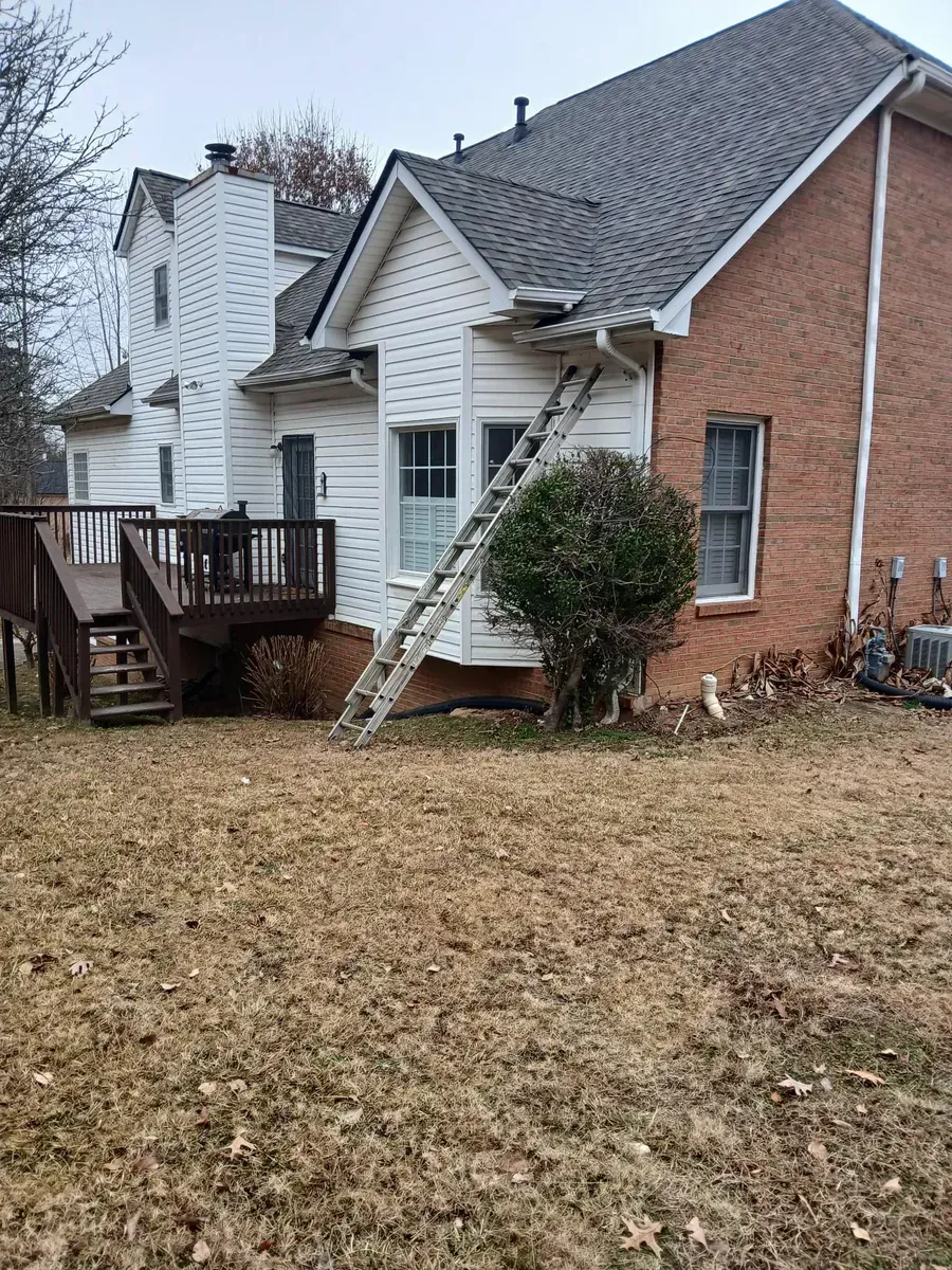 Backyard view of a house; ladder propped against the white siding near a window; brown deck and dry grass.