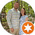 Couple smiling outdoors in front of trees. Man in plaid shirt, woman in blue floral dress.