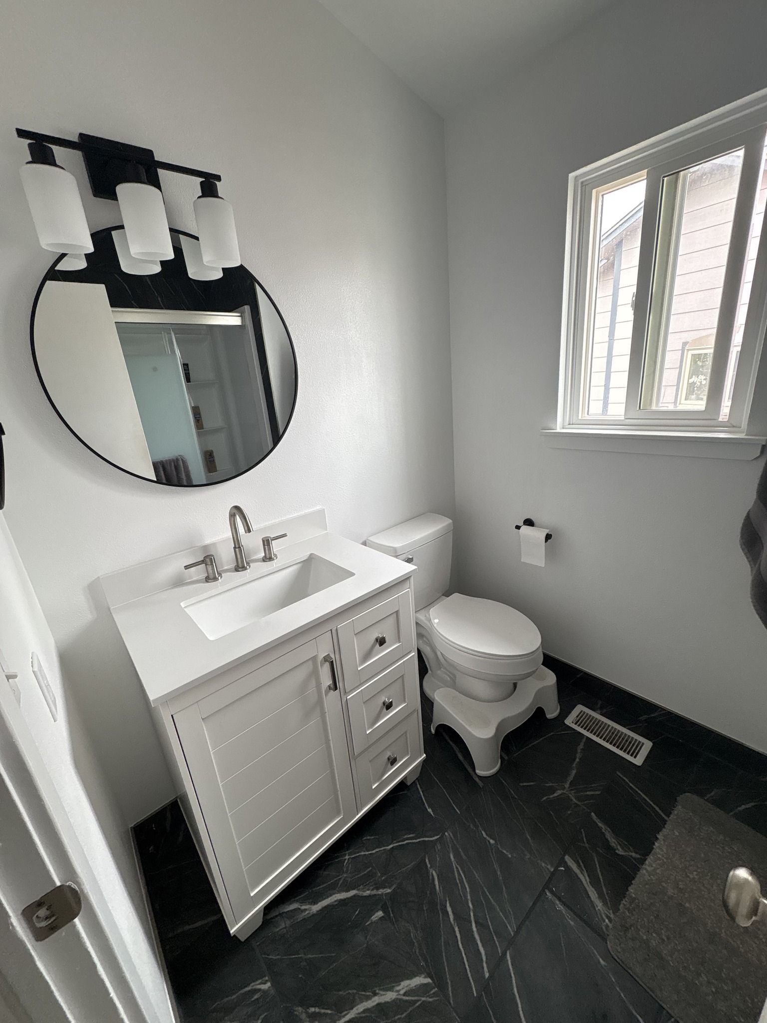 Small modern bathroom with white vanity, toilet, round mirror, black floor, and window.