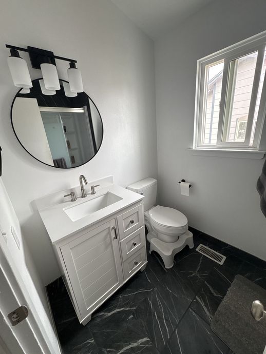 Small modern bathroom with white vanity, toilet, round mirror, black floor, and window.