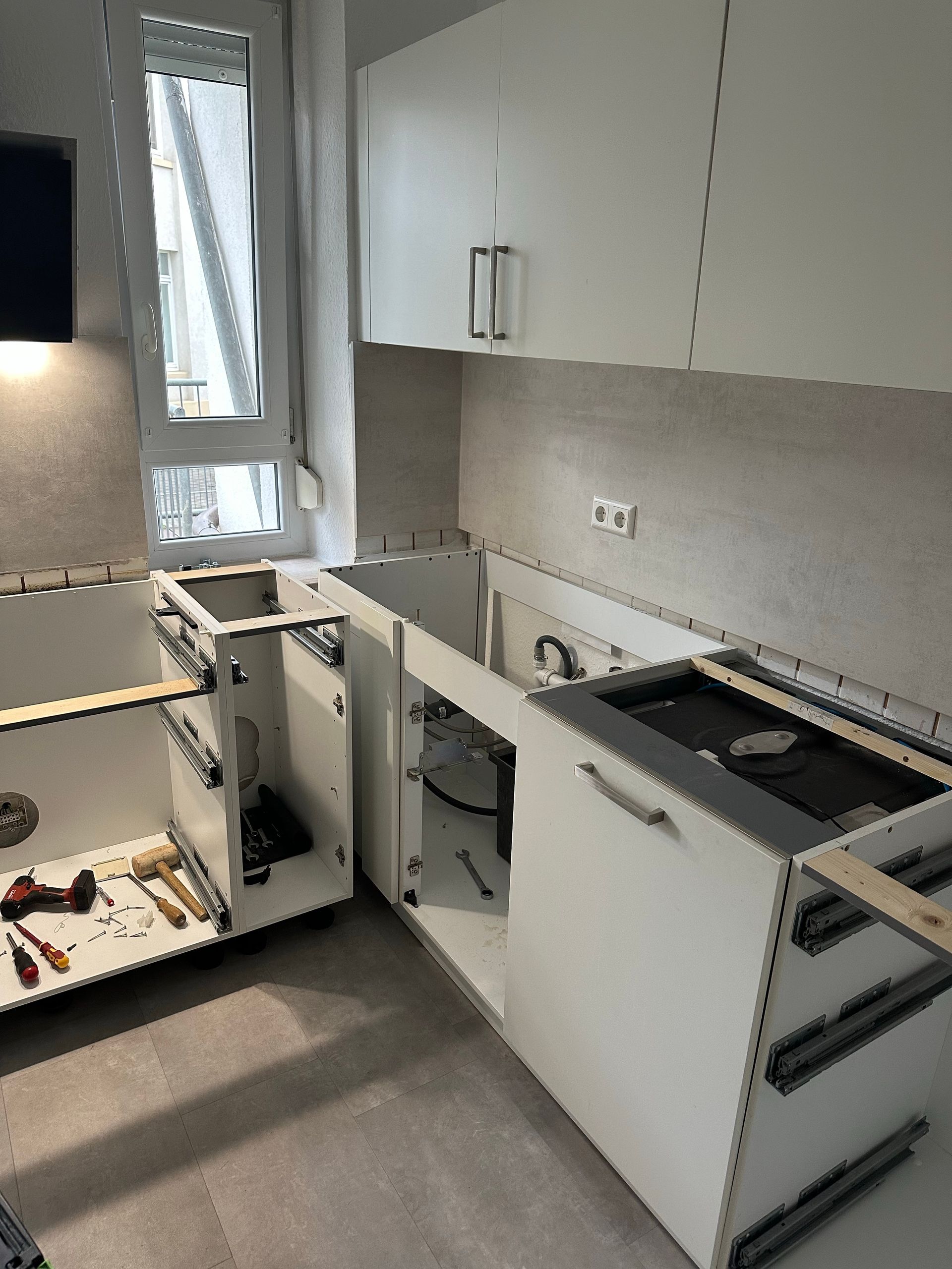 Damaged kitchen with open cabinets and drawers, tools on the floor, and a window above the sink.