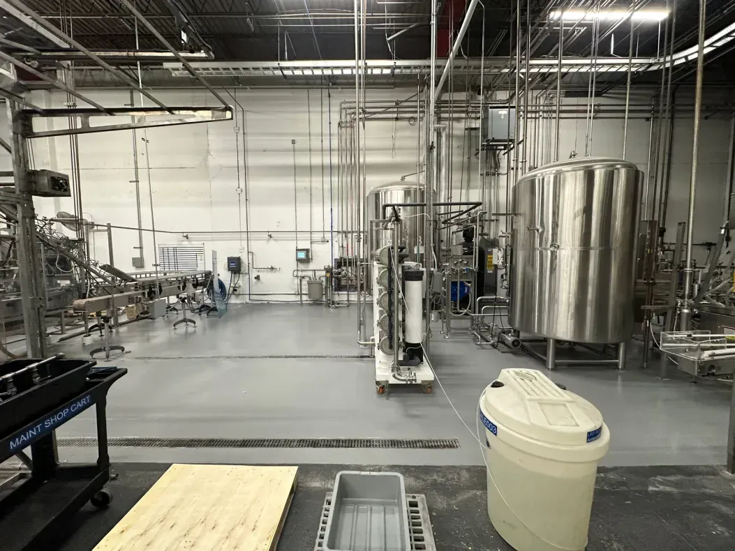 Industrial facility interior with stainless steel tanks, pipes, and equipment, and a light gray floor.