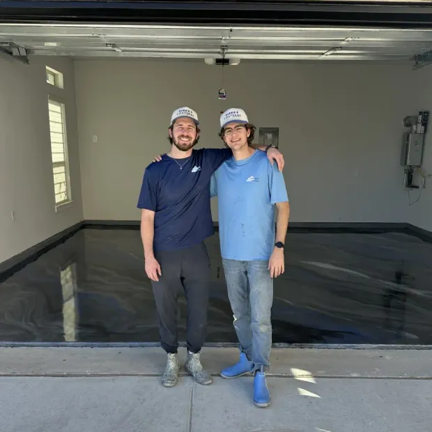 Two men standing in a garage, smiling with arms around each other, in front of a freshly coated black floor