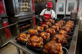 Rows of roasted chickens on trays in a commercial kitchen, with a worker in a red cap and white apron behind them
