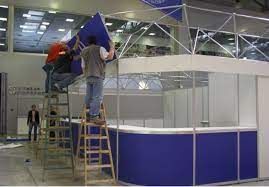 Workers on ladders installing a blue booth structure at an indoor trade show venue