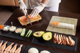 Hands slicing sushi rolls with gloved hands beside avocado, cucumber, shrimp, and dipping sauces on black trays