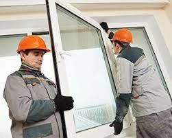 Two workers in orange hard hats carrying a large window frame indoors