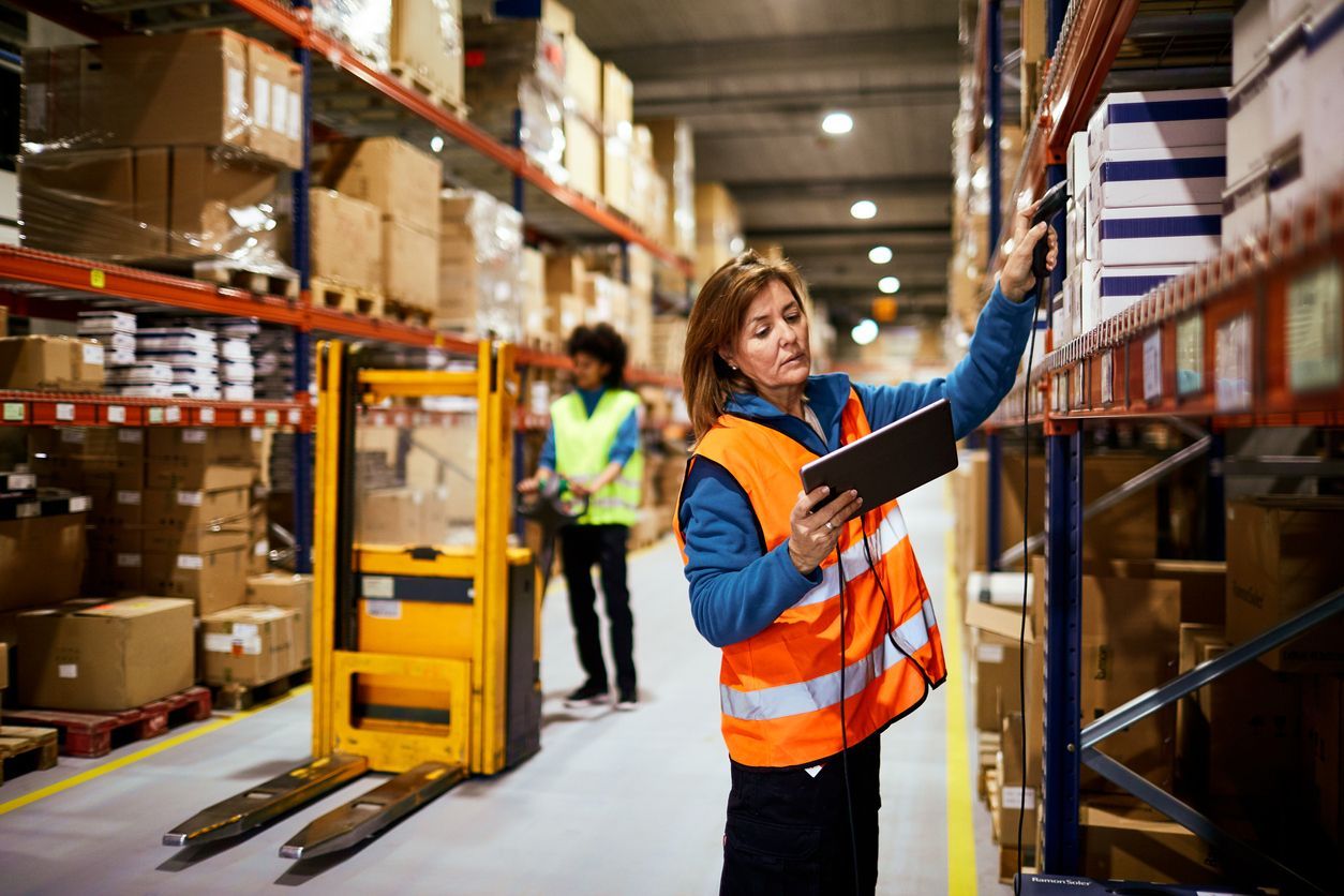 Warehouse worker in orange safety vest checking inventory on a shelf with a clipboard