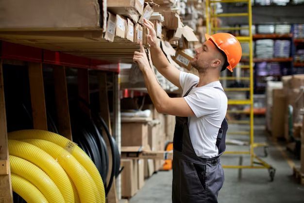 Warehouse worker in orange hard hat checking boxes on a high shelf beside coiled yellow hoses
