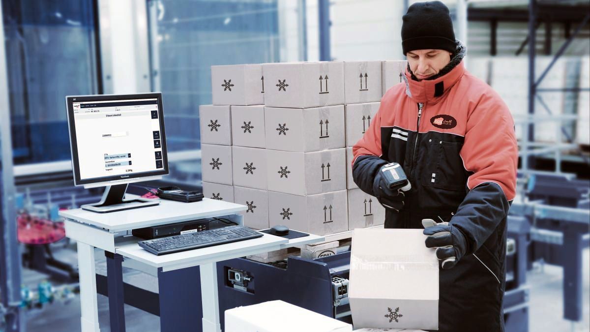 Worker in winter gear packing a cardboard box beside stacked boxes and a computer in a warehouse
