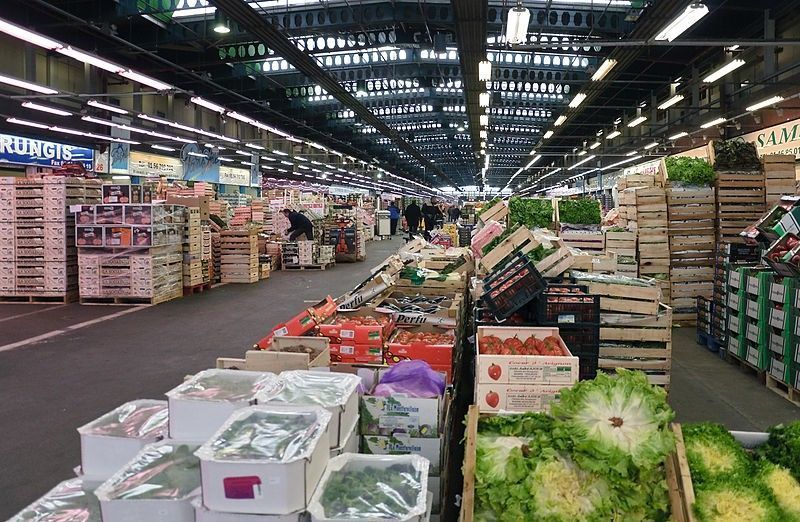 Produce section in a grocery warehouse with stacked crates and rows of fruits and vegetables under bright lights