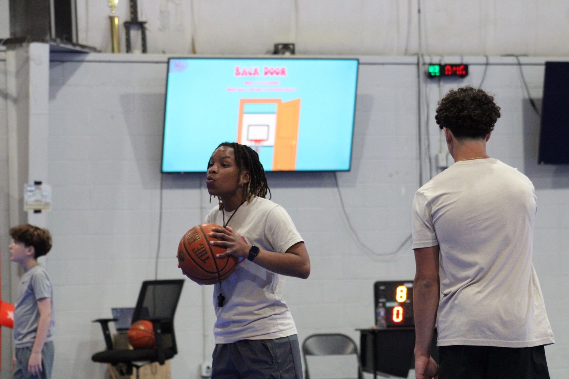 A coach holds a basketball in a gym near a scoreboard, facing a screen showing a basketball hoop diagram.