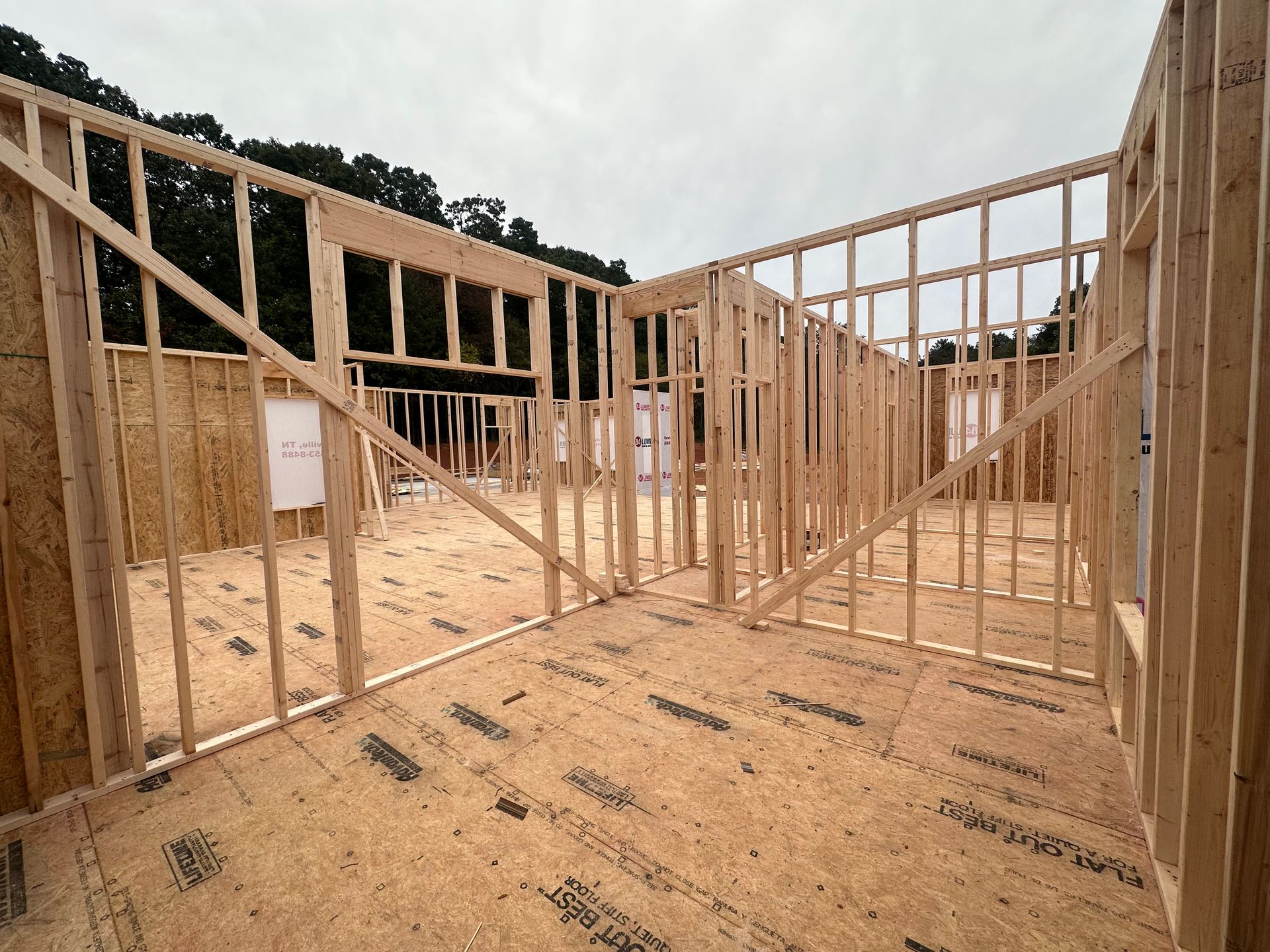 Wood-framed house under construction with open interior framing and plywood subfloor, viewed from inside.