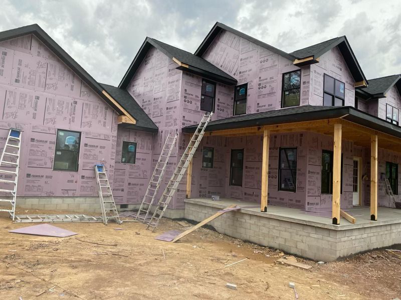 Two-story house under construction with white sheathing and a person on scaffolding under a clear blue sky