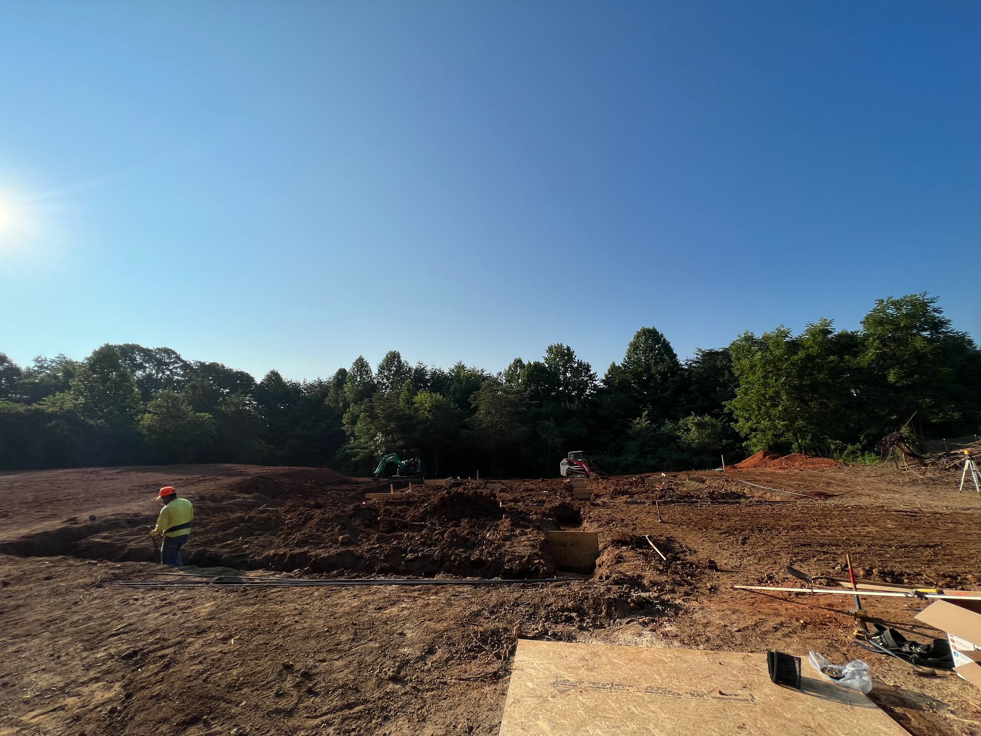 Wide dirt construction site with excavated earth, a worker in a yellow vest, and trees under a clear blue sky