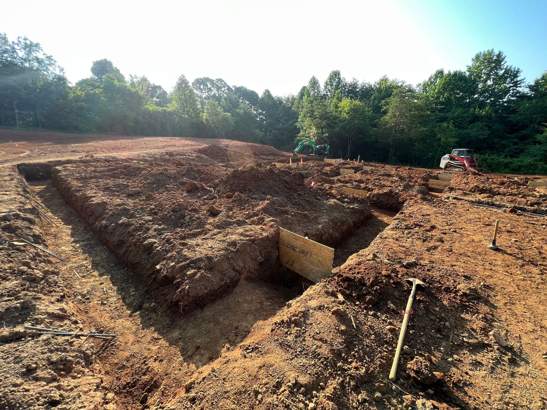 Excavated dirt pit in a clearing, with a wooden support panel and trees in the background