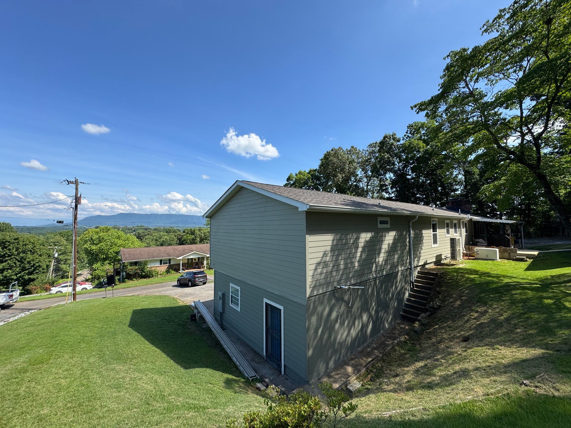 A green hillside house with a blue roof under a sunny sky, overlooking distant mountains.