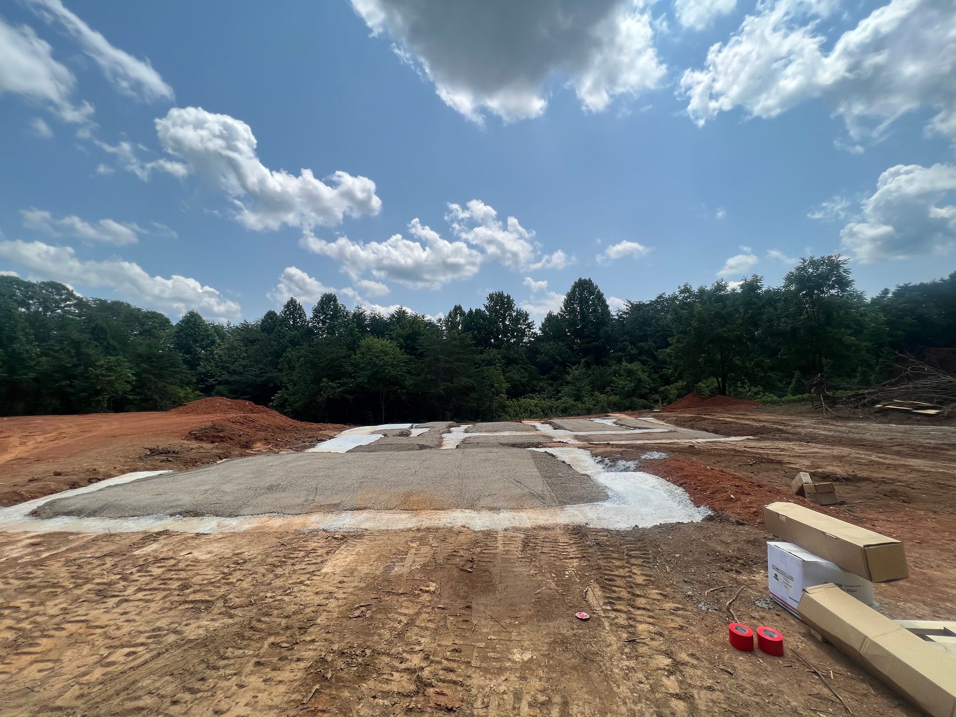 Cleared construction site with concrete slab, dirt foreground, trees, and cloudy blue sky