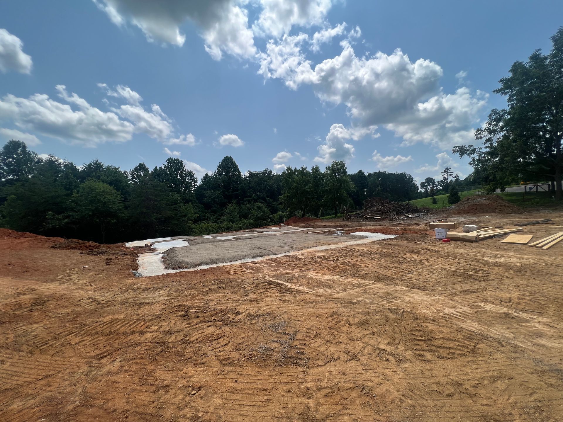 Cleared dirt lot with white concrete patches, trees in back, and a cloudy blue sky