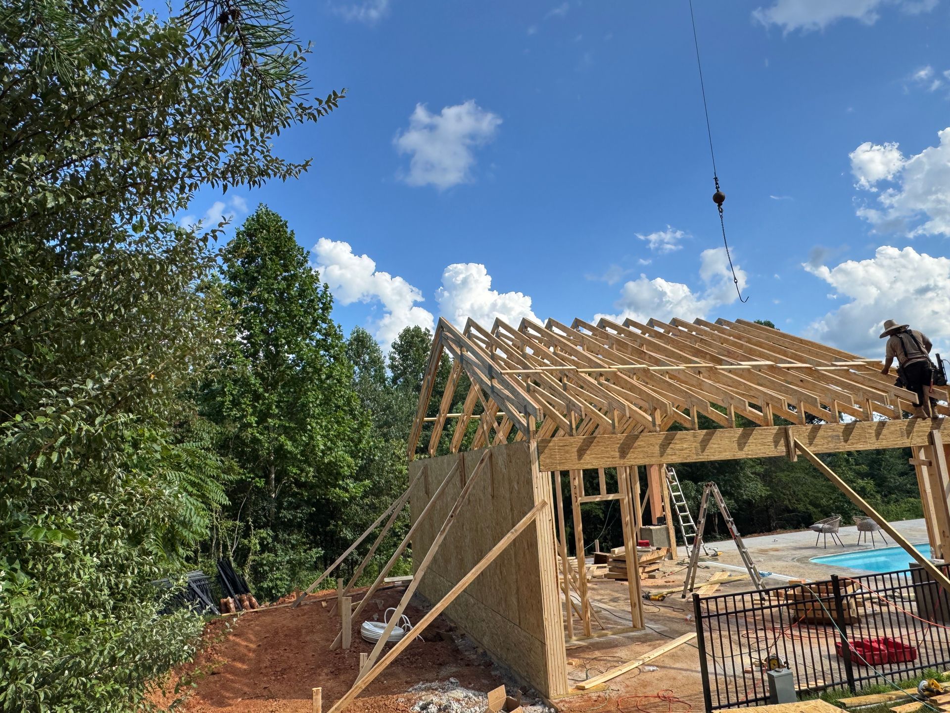 Partially built wooden house frame under construction on a sunny day, with worker on the roof and trees nearby