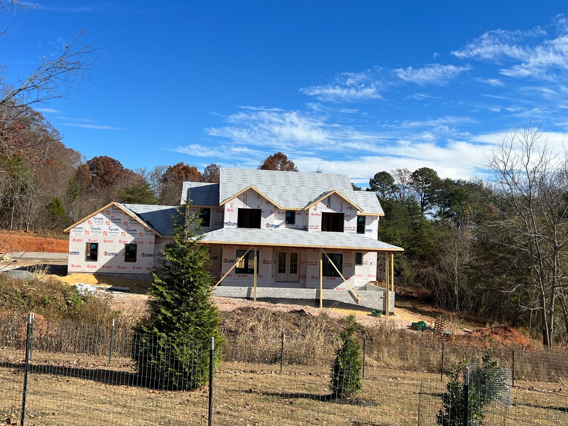 Unfinished two-story house with white siding and stone base on a wooded lot under a blue sky.