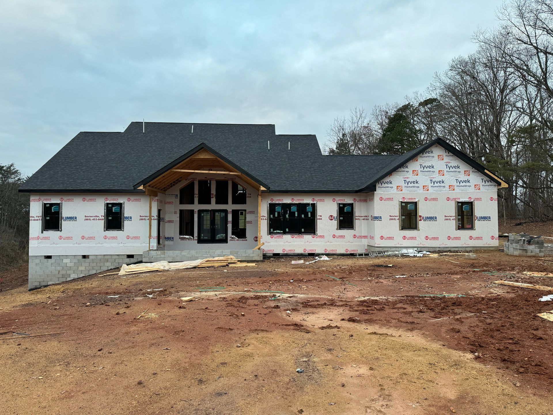 Unfinished single-story house under construction with white exterior and dark roof on bare dirt lot.