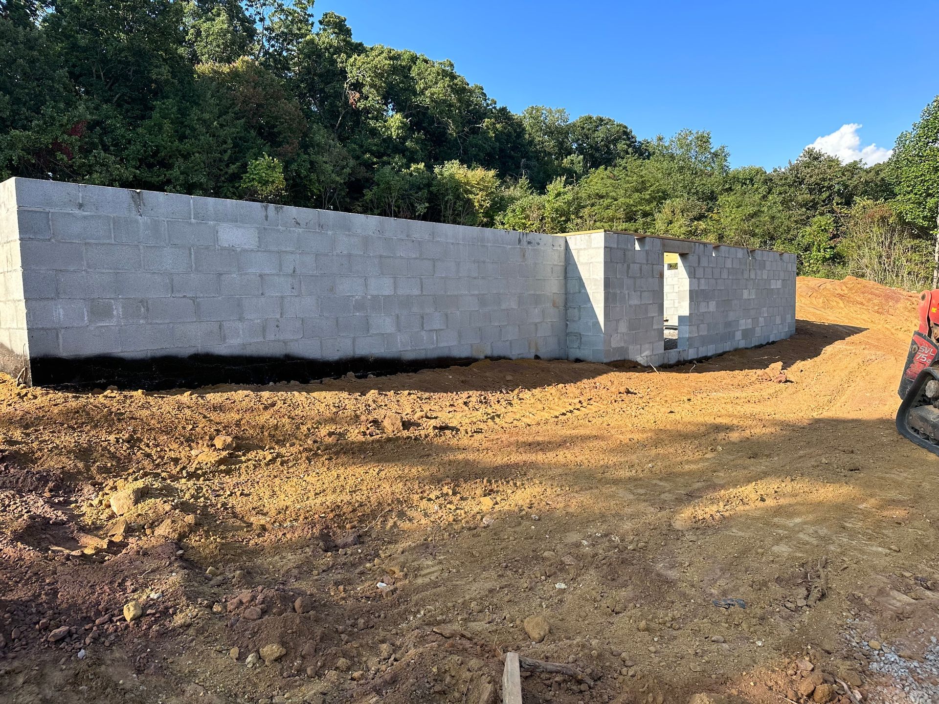 Partially built cinder block wall on a dirt lot with trees and blue sky in the background