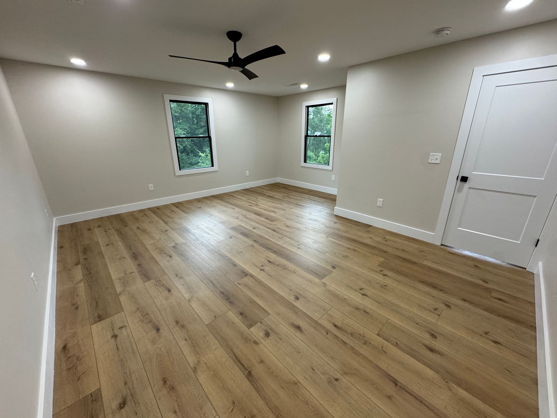 Empty beige room with wood floors, two windows, ceiling fan, and white door
