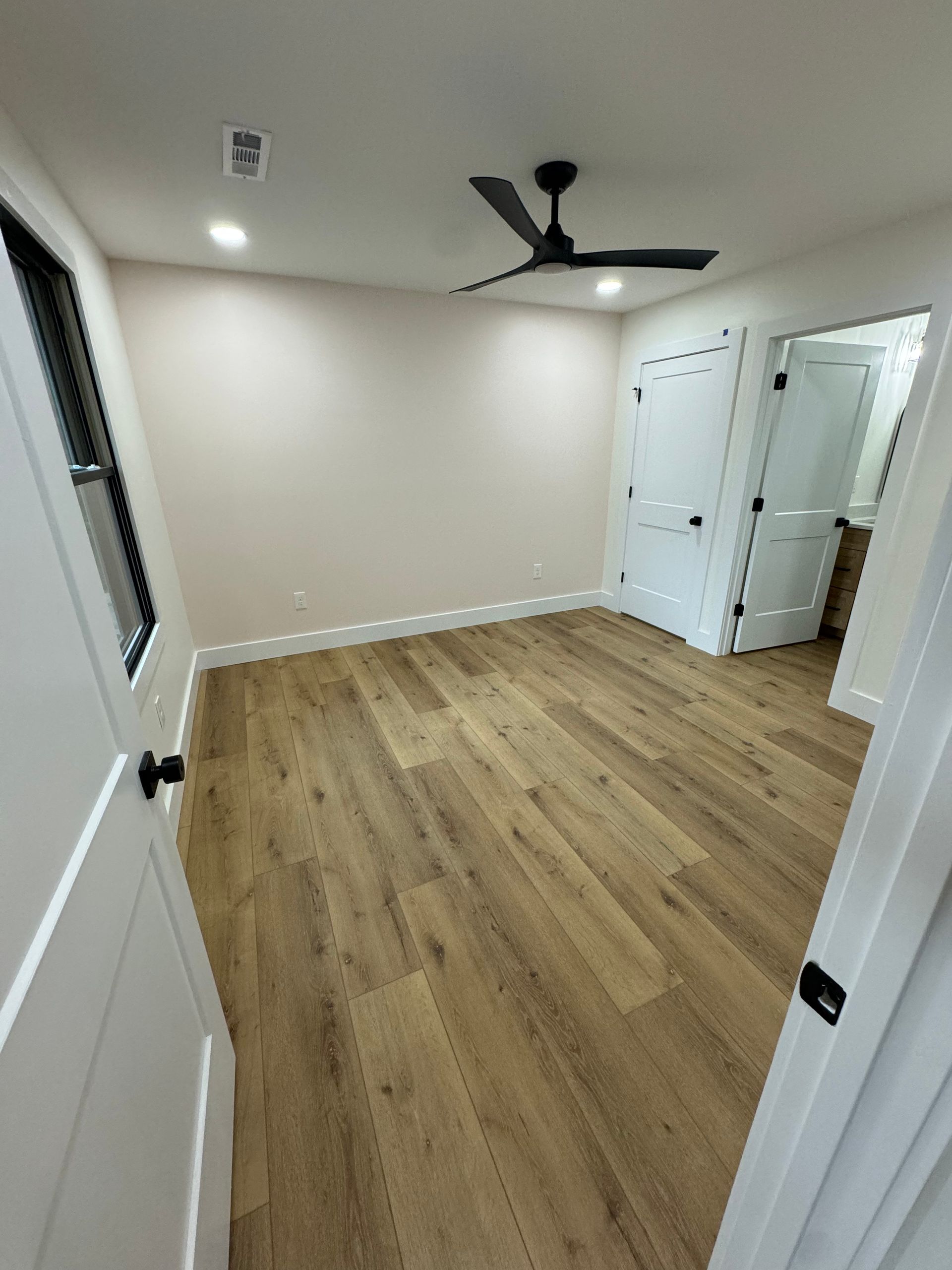 Empty beige bedroom with wood floors, white doors, and a black ceiling fan.