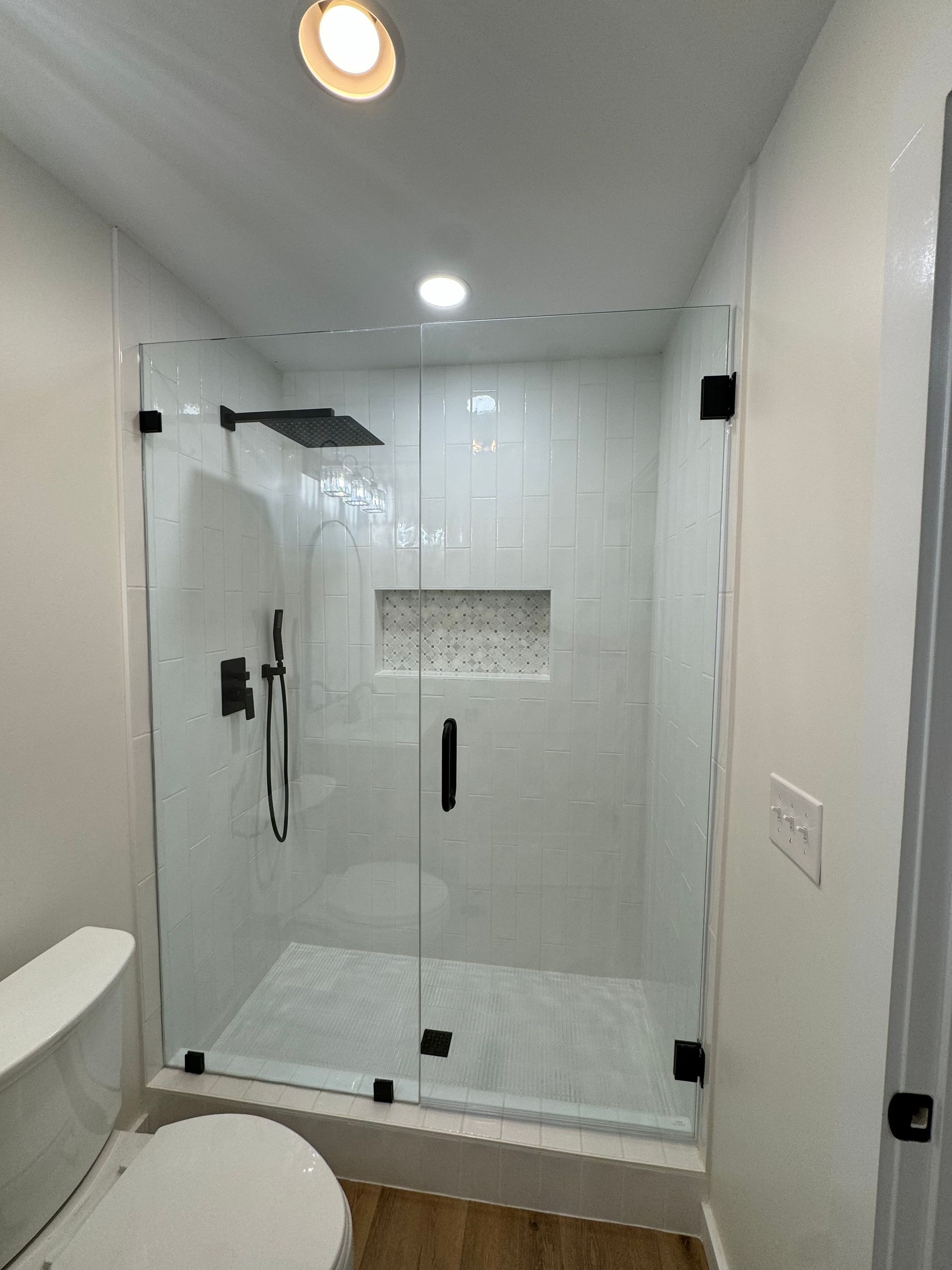 Modern white walk-in shower with glass door, built-in shelf, and recessed ceiling light in a narrow bathroom