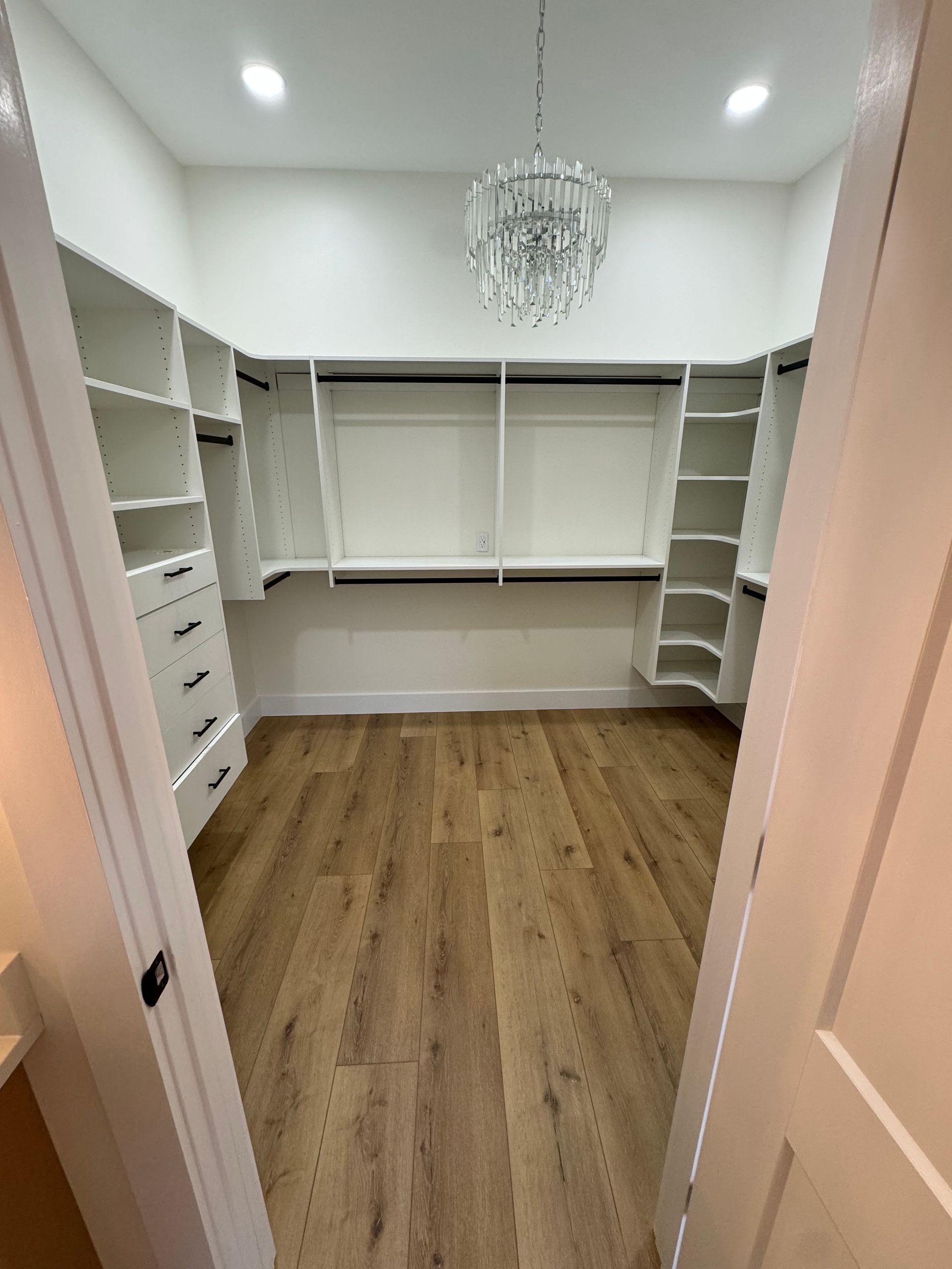 Walk-in closet with white built-in shelves and drawers, wood floor, and a chandelier overhead.