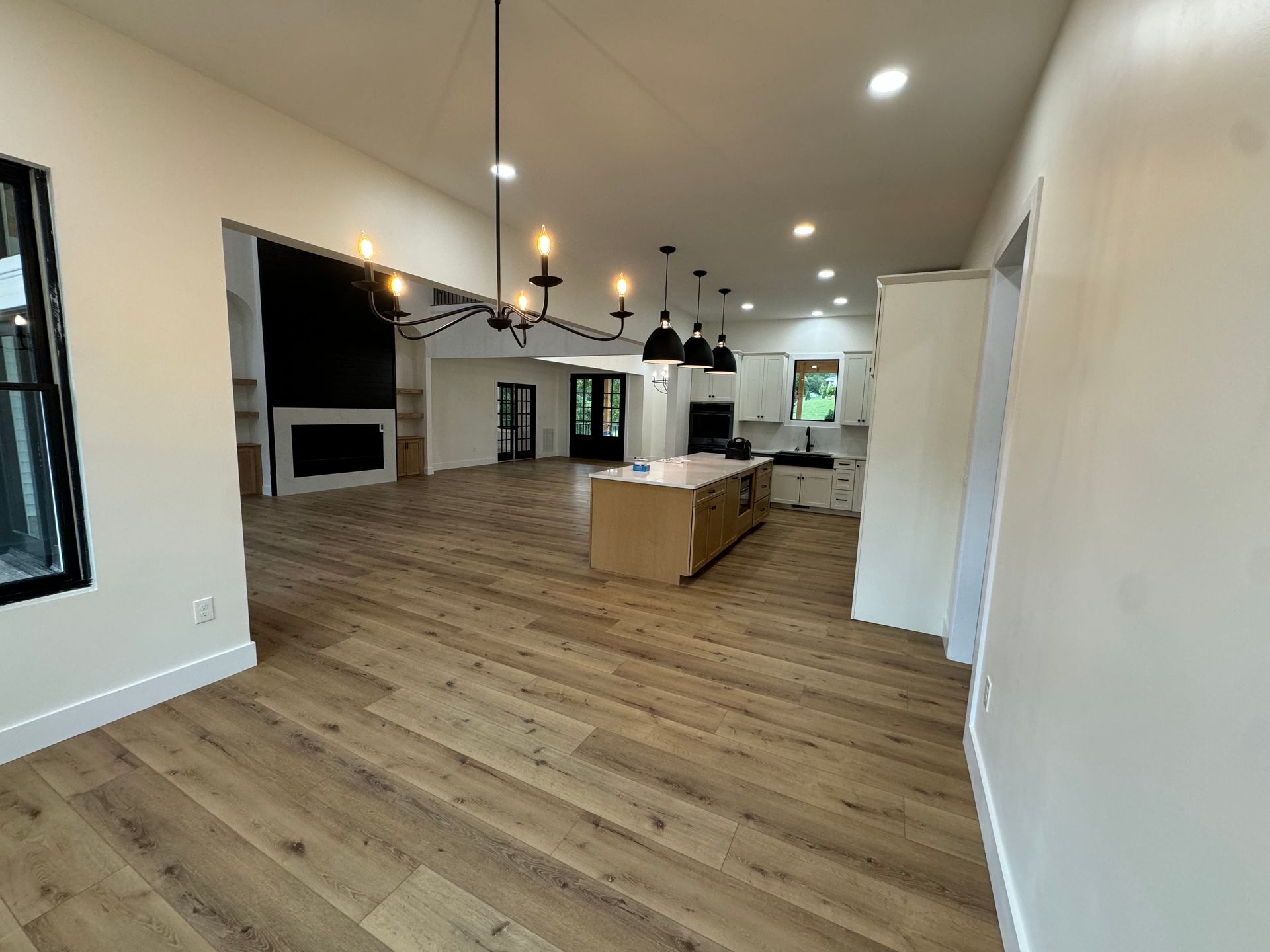 Empty modern living room with wood floors, white walls, black fireplace, and kitchen island in background