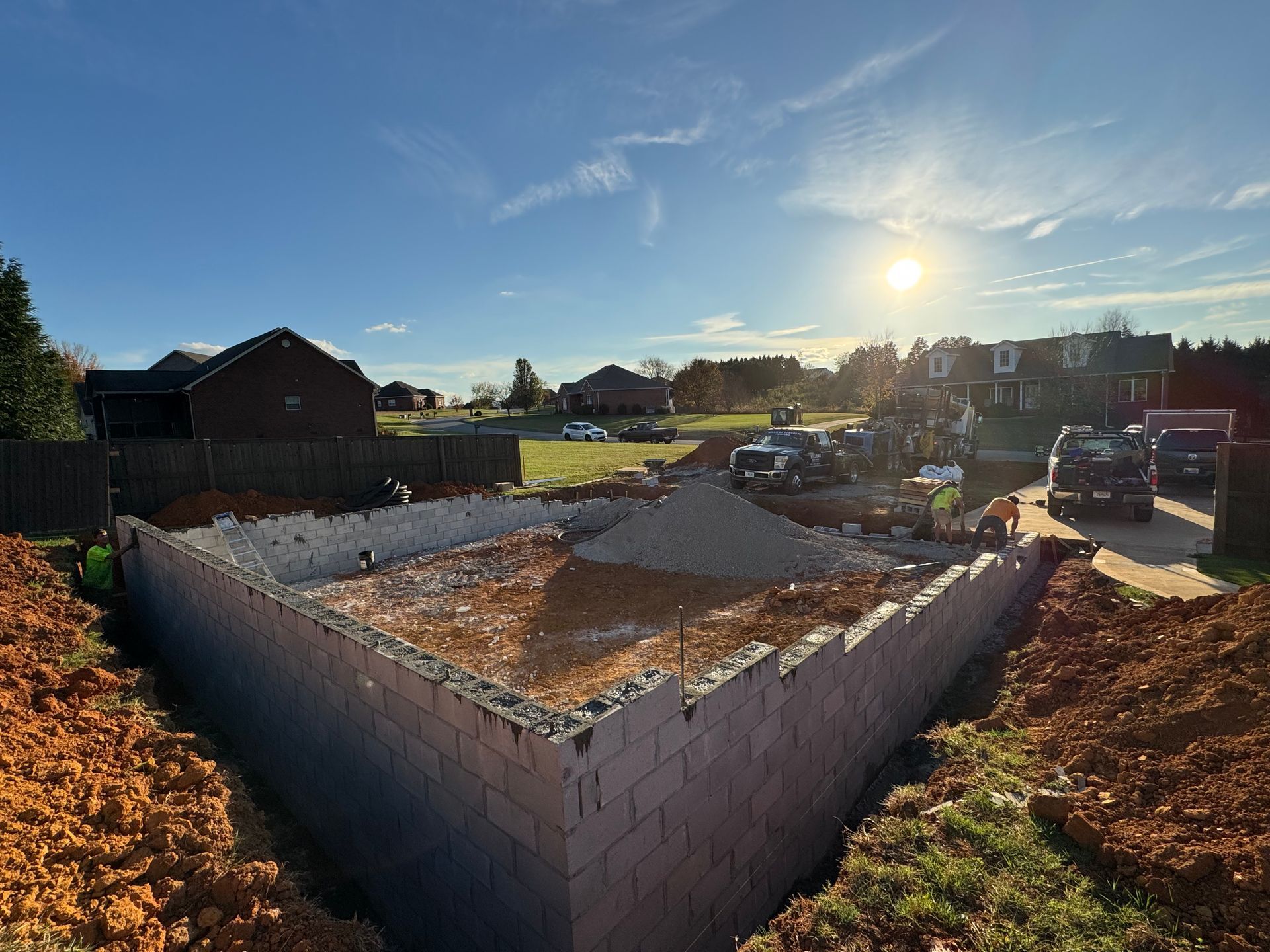 Sunlit construction site with a brick foundation, gravel inside, and houses in the background