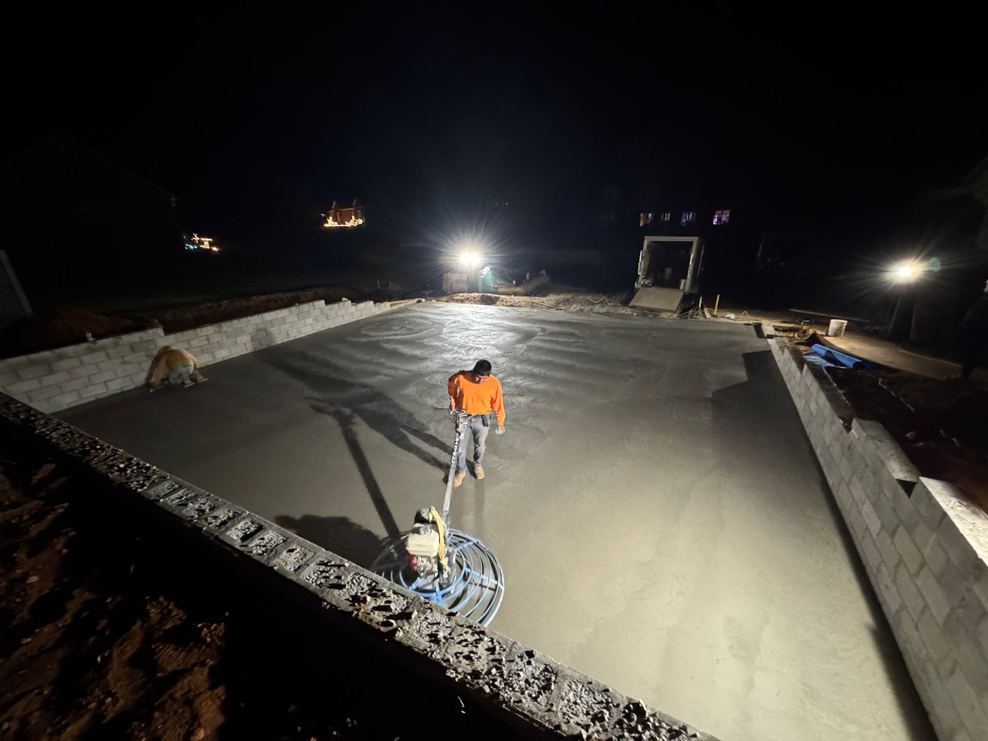 Worker in orange vest smoothing concrete at night on a large construction site.