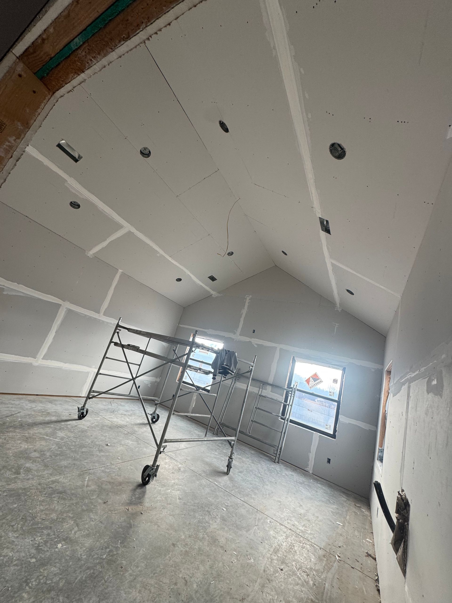 Empty room under construction with drywall, scaffolding, and concrete floor under bright window light