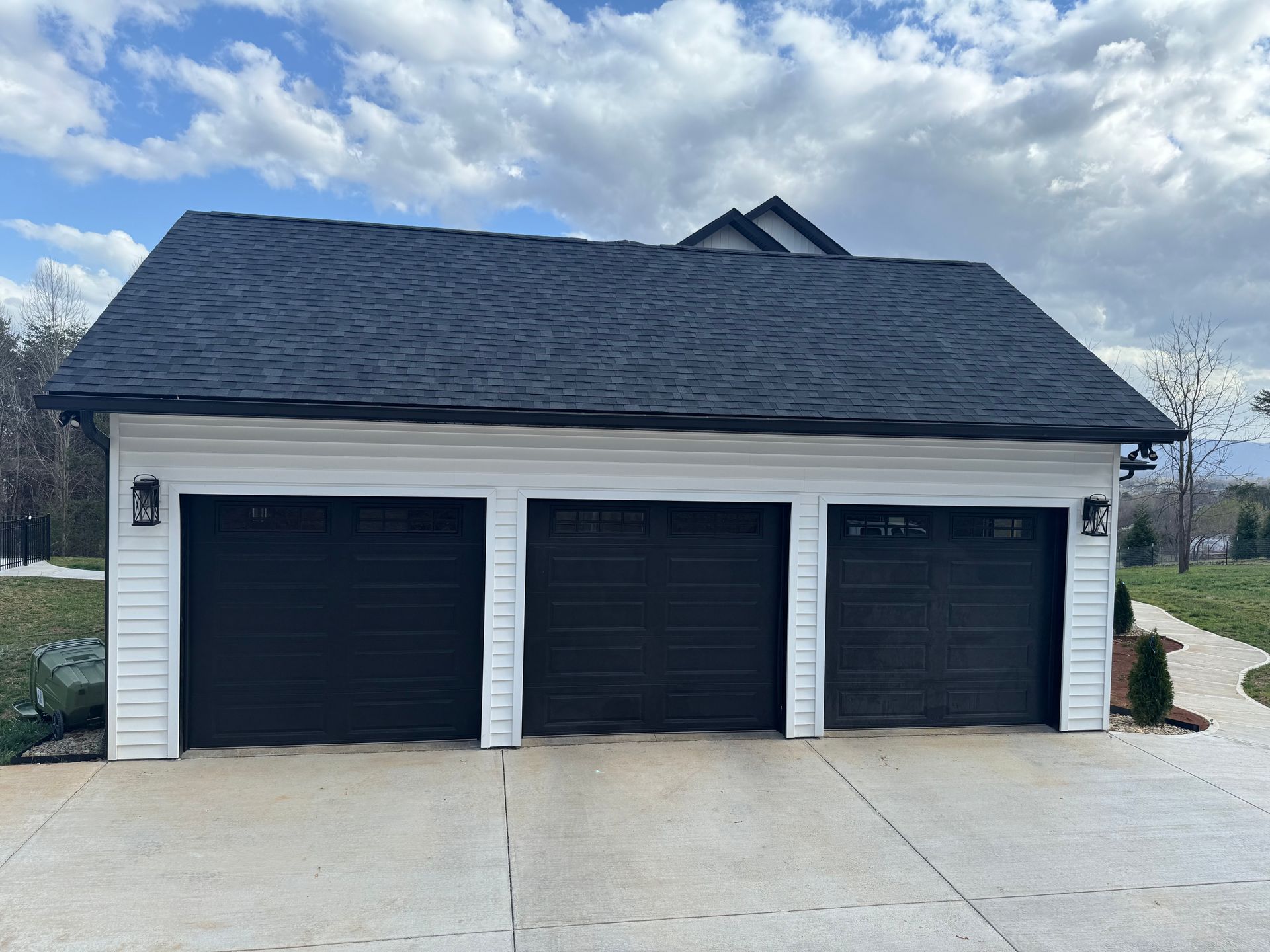 Three-car garage with white siding, black doors, and dark shingled roof under a cloudy sky